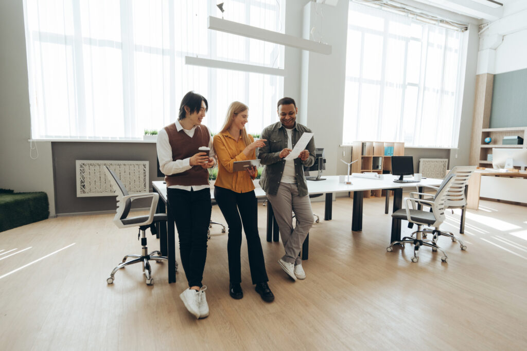 Three young colleagues of different ethnicities are standing in their office, discussing a project and smiling